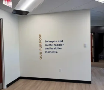 Office hallway with a white wall displaying vertical gold text 'OUR PURPOSE' and blue text 'To inspire and create happier and healthier moments.' Exit sign visible above.